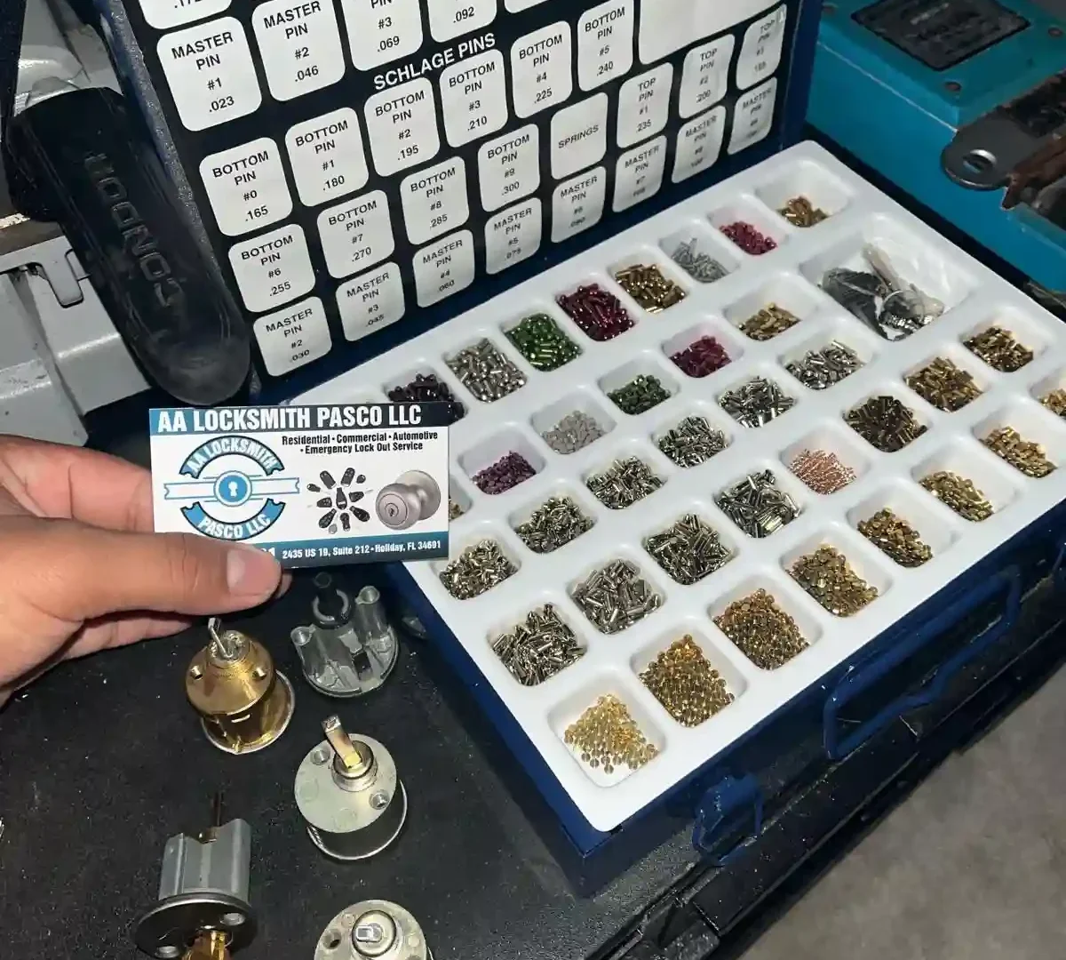 A person holding a locksmith rekeying pin kit for Kwikset and Schlage locks, showcasing labeled compartments of pins and springs, lock cylinders, and a business card.